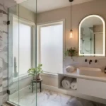 Modern renovated bathroom with large format marble-look tiles in shower, matte black fixtures, floating white vanity, arched backlit mirror, and open shelving with rolled linen towels — high-end bathroom on a budget